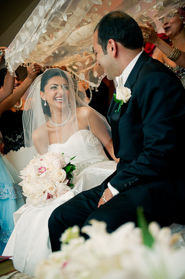 the happy bride and groom smiling and laughing at each other during traditional ceremony ritual - bride is wearing white dress with embellishments and full length veil while holding ivory, light pink, and green bouquet - photo by Houston based wedding photographer Adam Nyholt